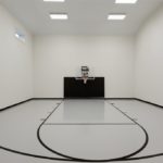 Millz House installed basketball court featuring white knight gray floor coating, black lines, black wall pads and 60" adjustable wall-mount hoop