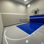 Multi-game court featuring floor coating in white knight gray with standard blue lane, white basketball lines, Gladiator 60" adjustable basketball hoop, royal blue wall pad, volleyball net, black cove base