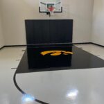 Indoor basketball court featuring floor coating in white knight gray with black lane, custom logo, Gladiator 60" adjustable basketball hoop, black wall pad, black cove base