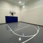 Indoor basketball court featuring floor coating in standard gray, white game lines, Gladiator 60" adjustable basketball hoop, royal blue wall pad, black cove base