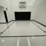 Indoor basketball court featuring floor coating in light gray, black game lines, Gladiator 60" adjustable basketball hoop, black wall pad, black corner pad, black cove base